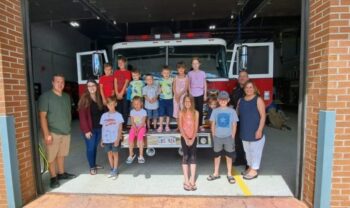 Douze enfants et trois adulte devant un camion de pompier ayant les deux portes aux extrémités ouvertes, devant une entrée de caserne de pompier en plein jour