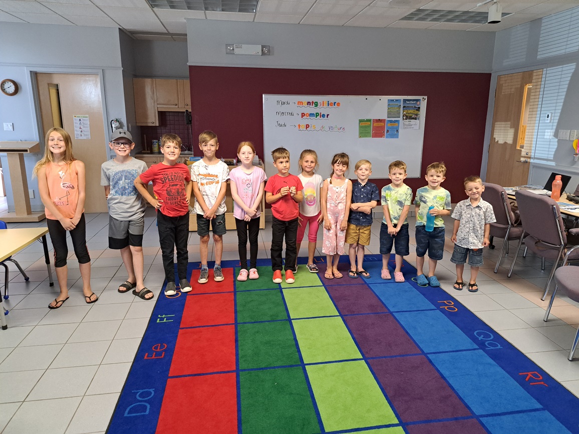 Une dizaine d'enfant en ordre de grandeur de gauche à droite, sur un tapis de carrés de couleurs diverses, dans la pièce d'une bibliothèque