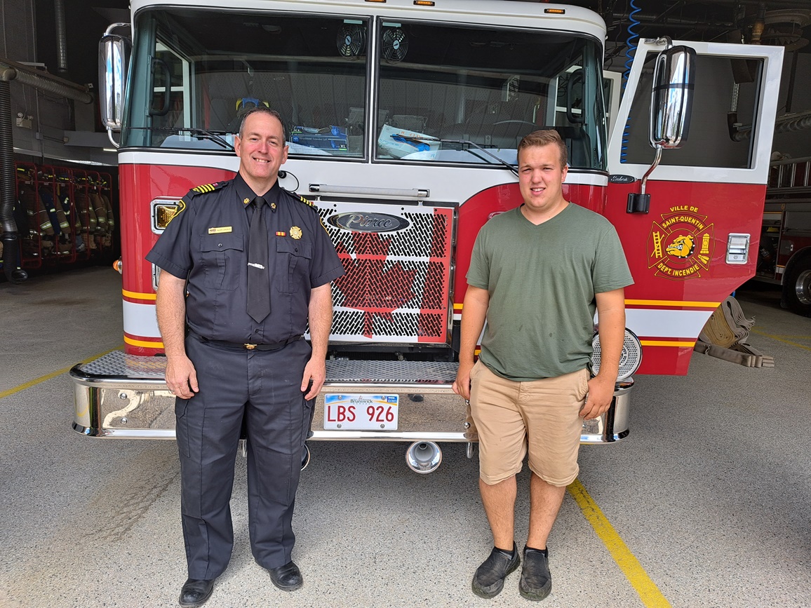 Un homme en uniforme de pompier et un autre homme habillé normalement devant un camion de pompier, dans un caserne de pompier