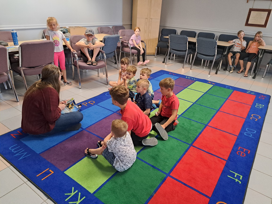 Une dizaine d'enfants sont assis leur animatrice du club de lecture. Cette dernière leur fait la lecture. Ils sont assis sur un tapis comportant plusieurs carrés de couleurs différentes