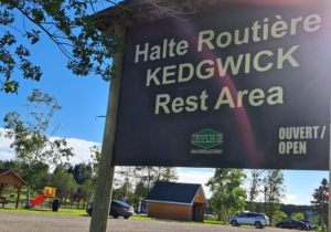 Pancarte affichant la halte routière de Kedgwick avec trois automobiles stationnées devant le parc et par une belle journée dégagée