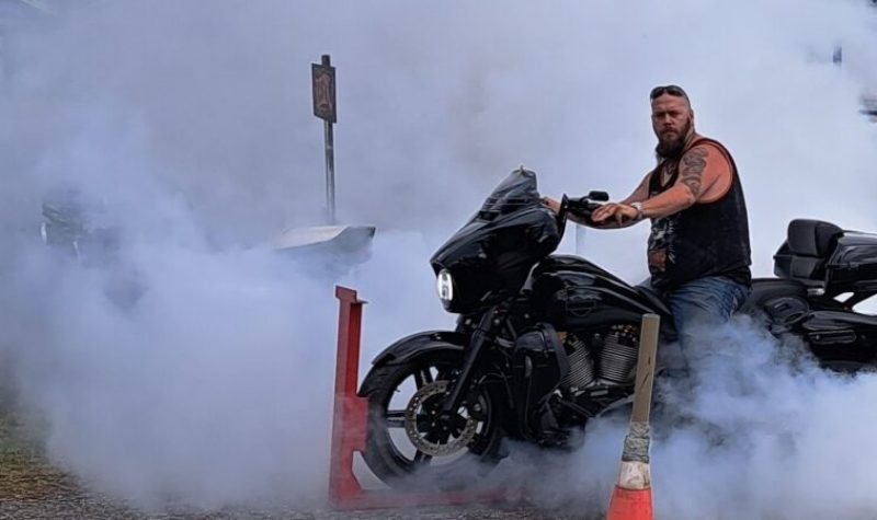 Un homme en camisole noir sur un moto noir entouré de boucane fait par le frottement de son pneu arrière sur du béton.