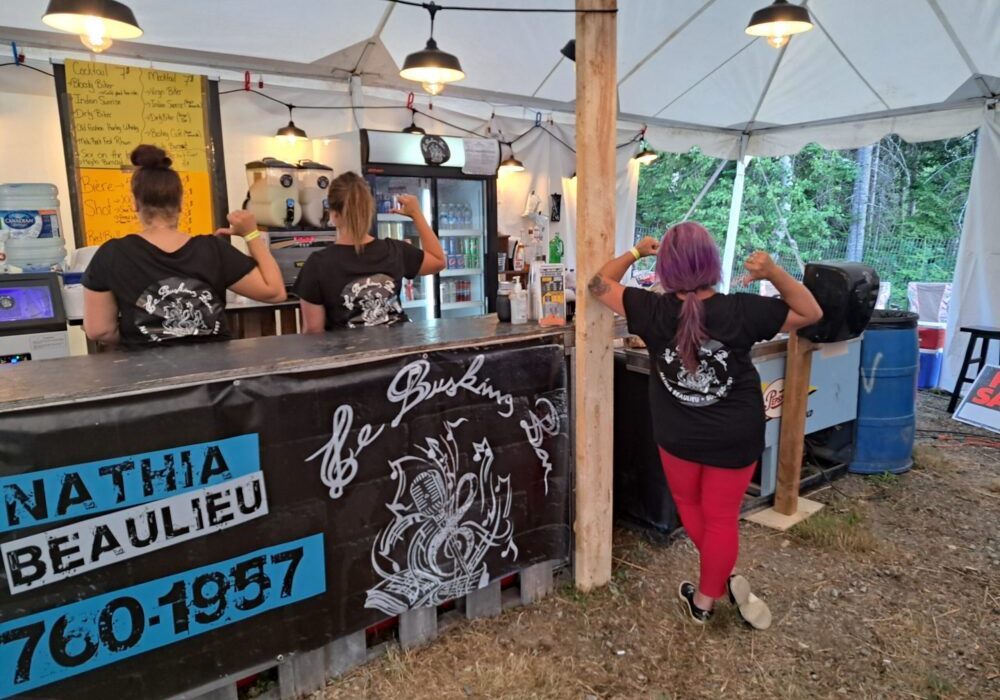 Trois femmes à dos tourné pointant vers le logo de leur chandail noir, derrière le bar d'un festival.