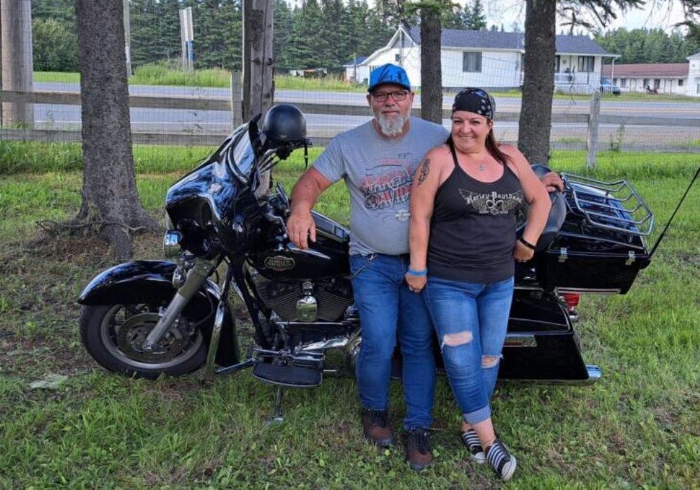 Couple côte-à-côte se tenant devant leur moto, devant des arbres et un motel en arrière plan