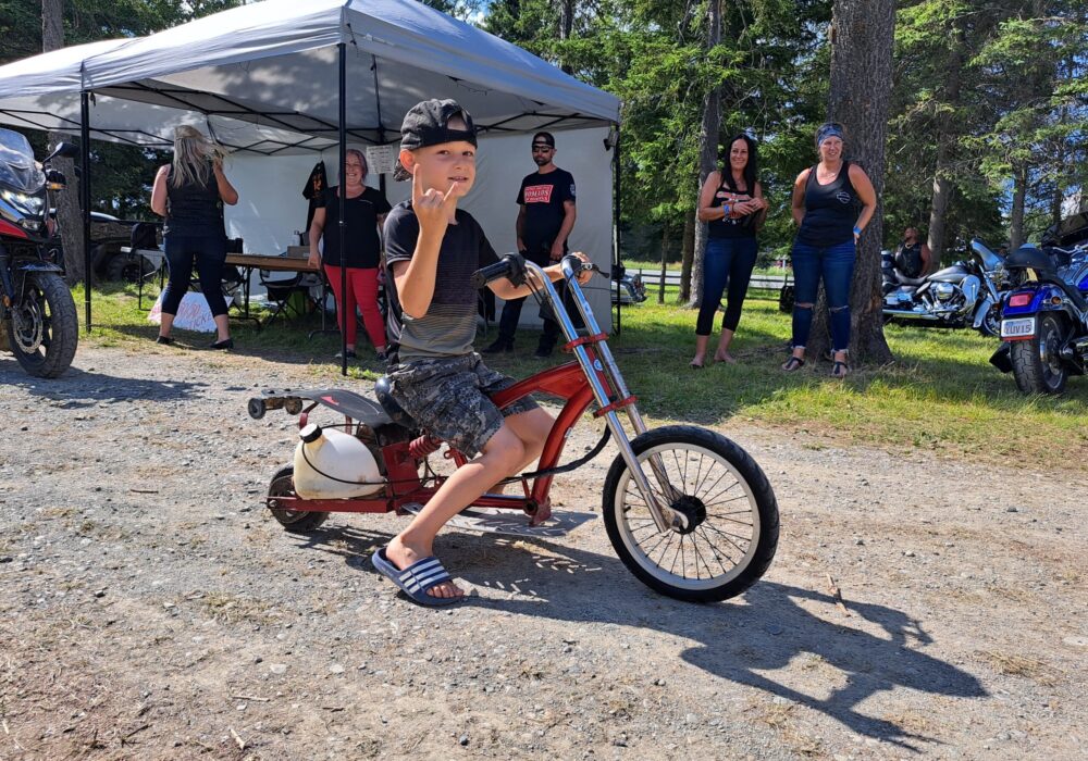 Jeune garçon sur une très petit motocyclette modifiée, une chemin en garnotte et sur le regard de quelques personnes qui sont derrières