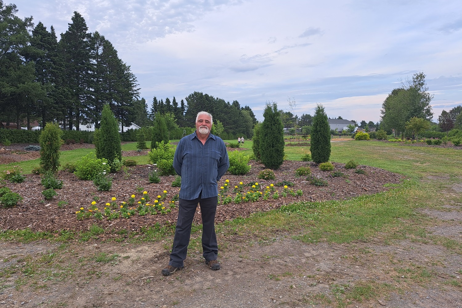 Homme portant une chemise bleue se tenant droit un parc comportant des arbres et des plantes lors d'une soirée nuageuse