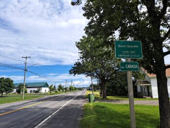 Une rue, des arbres, et un ciel bleu nuageux avec un panneau indiquant les limites de la Ville.