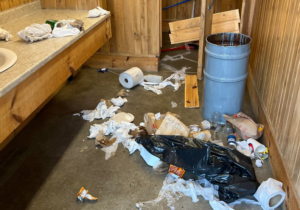 Salle de bain publique ayant subi des actes de vandalisme. Poubelles renversé, papier de toilette sur le sol et porte enfoncée.