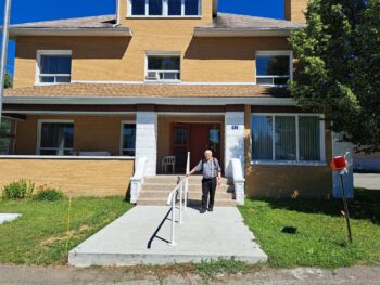 Presbytère de Kedgwick vu de l'extérieur. Le bâtiment est d'une beige avec quelques fenêtres. Une pelouse et un escalier en ciment sont devant. Un homme se trouve au pied de l'escalier.