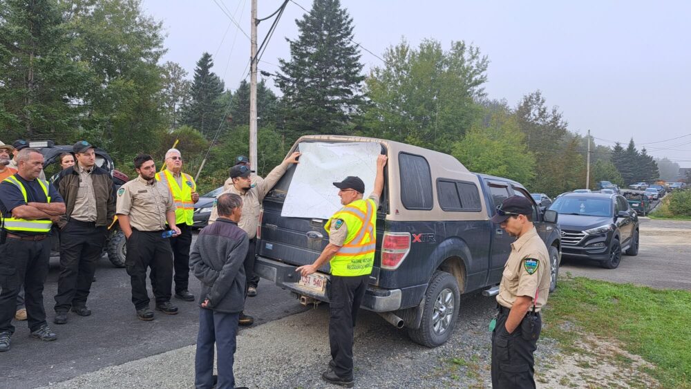 Homme portant un dossard de garde forestier tient une carte et explique à d'autres personnes devant lui la stratégie pour retrouver une personne disparue, lors d'un matin brumeux.