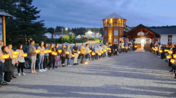 Plusieurs personnes en ligne tenant une lanterne lumineuse chacune, devant la tour des Pionniers à Kedgwick, alors que la noirceur tombe tranquillement.