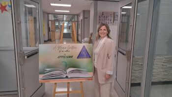 Une directrice d'école habillée proprement à côté d'une affiche à laquelle est inscrit le thème de son école : À l'école Marie-Gaétane, je me cultive en apprenant.