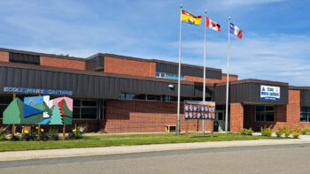 École en brique rouge et en tôle grise, affichant les drapeaux du Nouveau-Brunswick, du Canada et de l'Acadie.