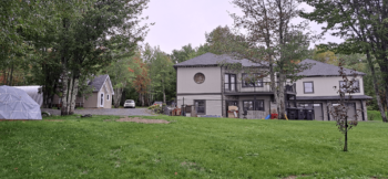 Une grande maison de couleur grise et mauve pâle autour d'une grand terrain de pelouse entouré d'arbres.