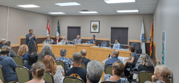 Homme parlant devant une assemblée ainsi que d'un conseil municipales, dans une salle rectangulaire bien éclairée.