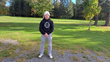 Homme avec une tuque blanche et un chandaille noire, debout devant un terrain de golf.