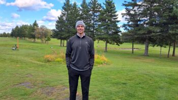 Homme vêtu de gris debout devant un terrain de golf dans une journée froide et ensoleillée.