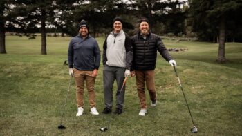 Trois hommes tenant leur bâton de golf sur un terrain de golf, lors d'une journée nuageuse et venteuse.