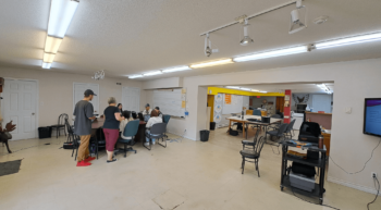 Une grande salle peinturée en blanc avec des élèves autour d'une table qui travaillent sur leur ordinateur.