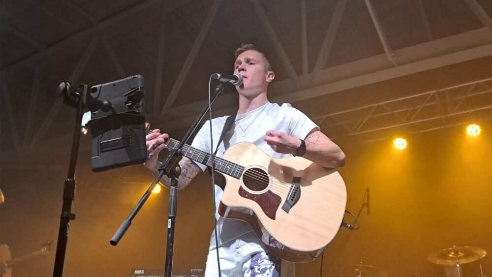 Homme vêtu de blanc tenant une guitare et chantant lors d'un concert.