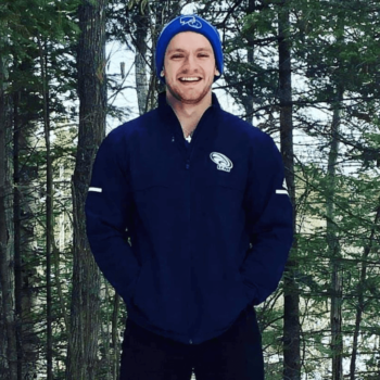Homme avec un manteau bleu foncé et une tuque bleu claire. Devant des arbres et souriant pour une photo.