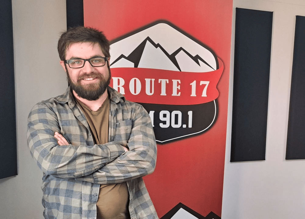 Homme portant une chemise et une barbe les bras croisés et souriant pour une photo devant inscrit Route 17 en blanc, rouge et noir.