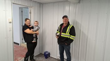 Une femme tenant un bébé au côté d'une homme portant un dossard, regardent l'appareil photo à l'intérieur d'une salle tout en blanc.