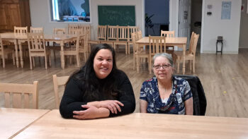 Deux femmes assises à une table et regarde l'appareil photo en souriant, dans une grande salle comportant plusieurs autres table identiques de couleur beige et en bois.