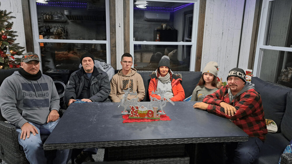 Six personnes sont assises autour d'une table noire. Derrière eux des fenêtres et un sapin de noël sur la gauche.