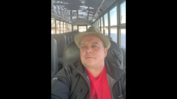 Chauffeur d'autobus au volant de celui-ci. Il porte un chapeau, une chemise rouge et une veste noire.