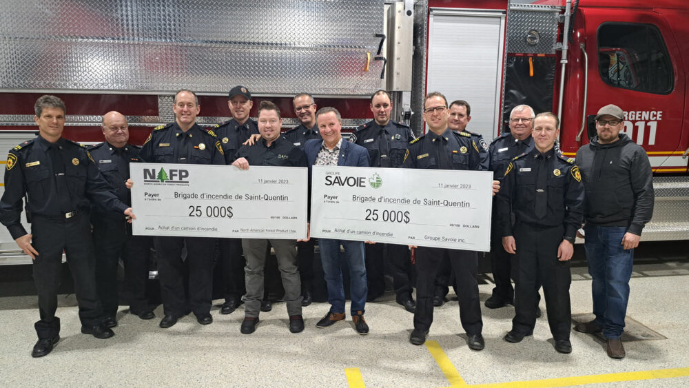 13 hommes en uniformes se tenant derrière 2 chèques format géant de 25 000 dollars respectivement. Derrière eux se trouve un camion de pompier. À l'intérieur d'une caserne de pompier.