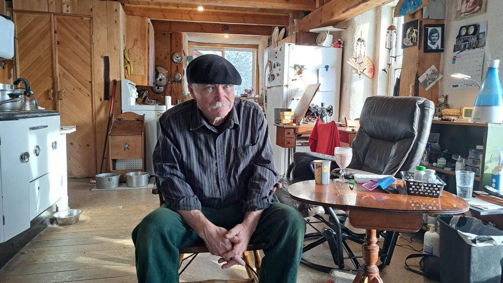 Homme portant un chemise et des pantalons assis dans sa maison portant un berret.