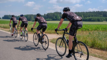Quatre cyclistes portant le même habit roule sur l'accotement d'une route, lors d'une belle journée ensoleillé avec quelques nuages.