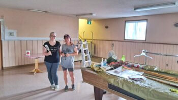 Deux femmes à l'intérieur d'une salle tiennent un pinceau à la main et s'apprêtent à peinturer la salle devant un table de billard contenant plusieurs outils de peinture.