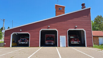 Caserne de pompiers de St-Jean-Baptiste-de-Restigouche. Trois portes de garages ouvertes avec trois camions de pompiers à l'intérieur. vue de face.