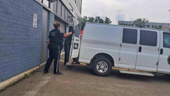Deux policiers en uniforme noir referme la porte arrière d'une camionnette blanche, lors d'une journée chaude et nuageuse.