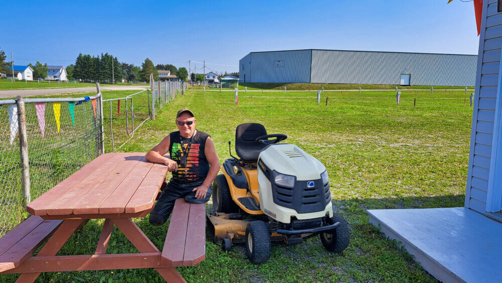 Un homme amputé des deux jambes est assis sur une table de pique nique, au côté de son tracteur à pelouse, sur un vaste terrain de pelouse, lors d'une journée chaude et ensoleillée.