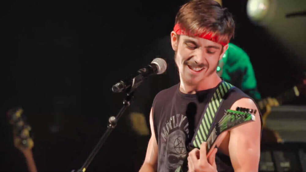 Un jeune guitariste et chanteur en action lors d'une spectacle. Il porte une camisole noire et un bandeau rouge sur la tête.