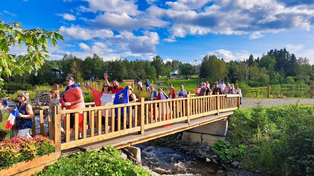 Un tintamarre aux couleurs acadiennes lors d'une soirée ensoleillée. Les gens marchent en traversant sur un pont une petit rivière.