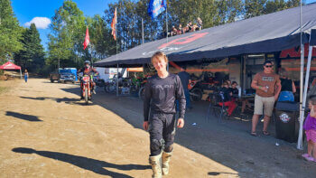 Un garçon de 15 ans debout en habit de motocross noir, lors d'une compétition de motocross pendant une journée ensoleillée.