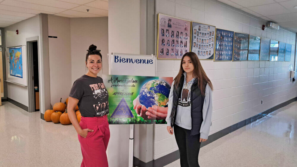 De gauche à droite, une enseignante et une élève devant l'affiche dévoilant la thématique de l'année scolaire 2023 : À Marie-Gaétane, j'explore le monde en apprenant.