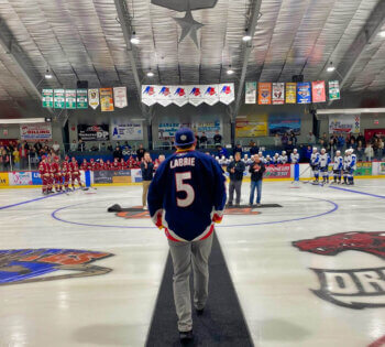 Mise en jeu protocolaire avant le début d'une partie d'hockey. Un homme fait dos à la caméra et marche sur un tapis vers le centre de la patinoire.