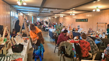 À l'intérieur d'un marché d'artisans où ceux-ci sont déguisé pour l'Halloween. une famille est assit autour d'une table et des visiteurs se promènent.