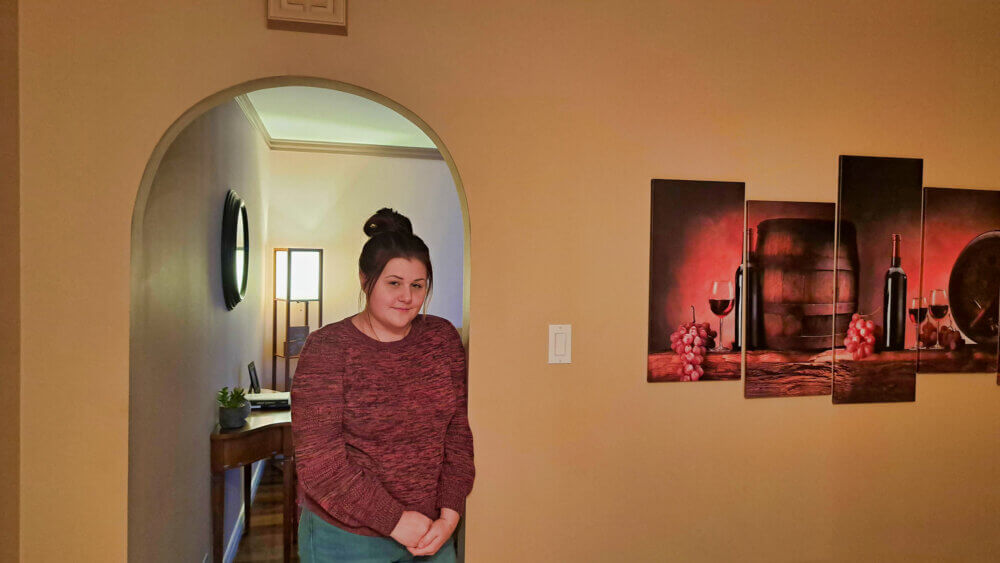 Femme vêtue d'un chandail rouge devant un mur pâle où est accrochée une toile aux couleurs rouge foncé.