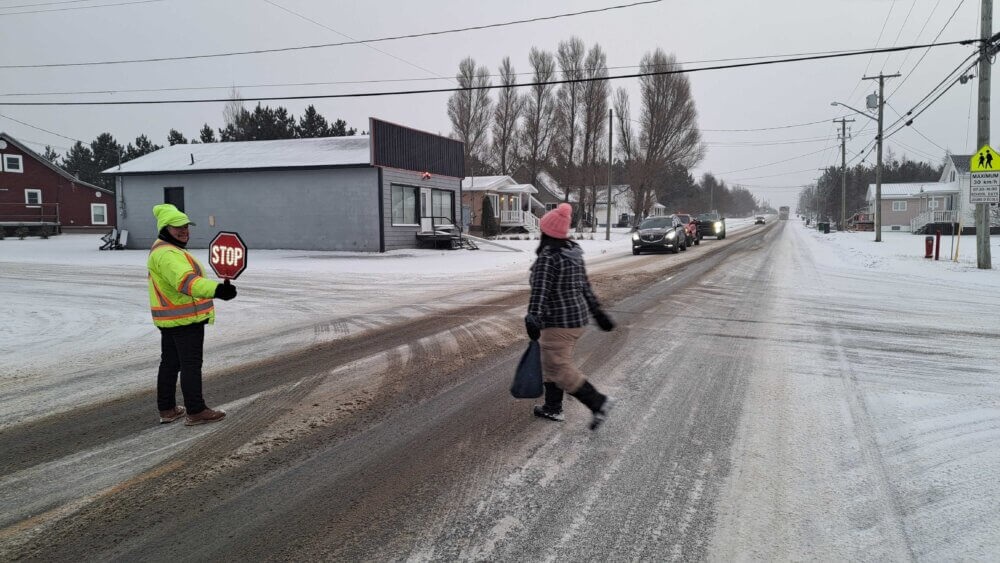 Une brigadière tenant une pancarte d'arrête aidant une écolière à traverser une rue enneigée. Des véhicules sont arrêtés pour laisser passer l'écolière, lors d'une journée de novembre nuageuse et enneigée