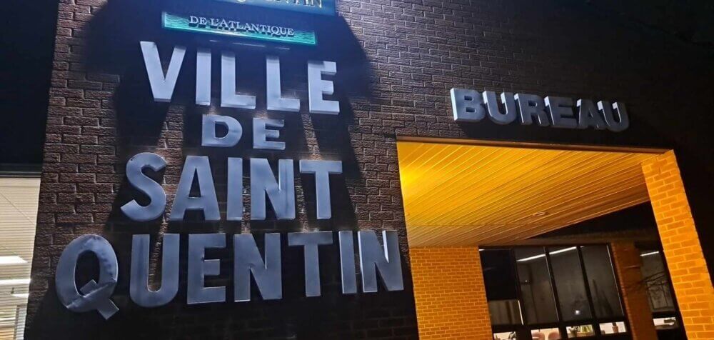 Le devant du bureau municipal de Saint-Quentin. Il fait noir à l'extérieur. Une lumière blanche éclaire les écrits "Ville de Saint-Quentin" à gauche et une lumière orange éclaire l'entrée principale de l'édifice.
