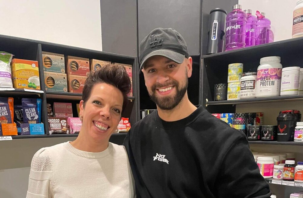 De gauche à droite, une femme au chandail blanc et un homme un chandail noir portant une casquette, les deux souriant pour une photo