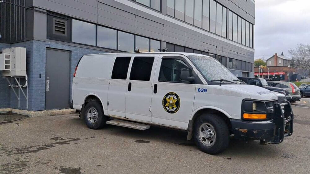 Camion de justice sécurité blanc devant un bâtiment foncé comportement plusieurs fenêtres de forme carré.