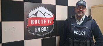 Le sergent Yannick Gagnon, vêtu d'un uniforme de police noire , à côté du logo de la Radio des Hauts-Plateaux indiqué " Route 17 "
