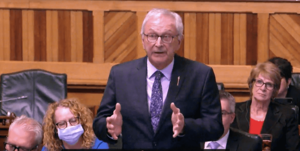 Le premier ministre du Nouveau-Brunswick, Blaine Higgs, à l'assemblée législative, vêtu d'un habit noir et d'une cravate bleue devant des membres de son parti politique